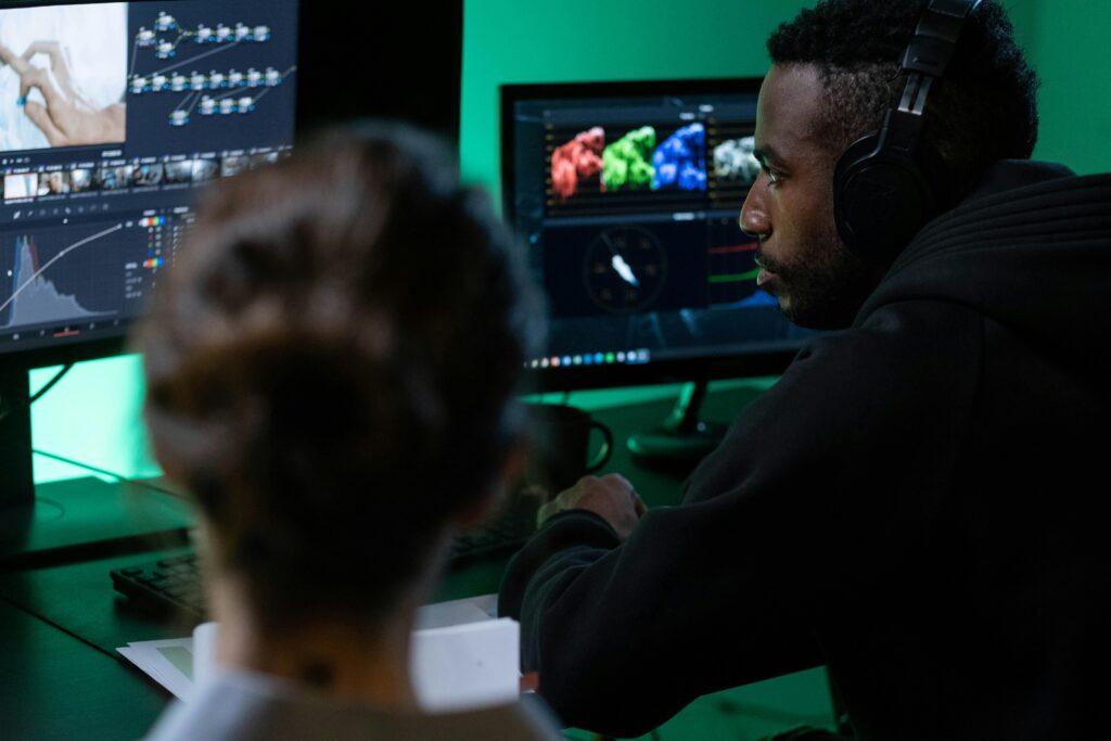 A man wearing headphones works on video editing with dual monitors in a dimly lit room.